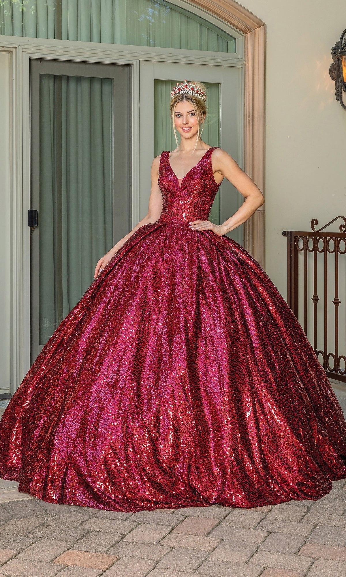 A woman stands outside in front of glass doors and a window, wearing the Quinceanera Dress 1558 by Dancing Queen—a sparkling, deep red ball gown with sequin details—paired with a jeweled crown.